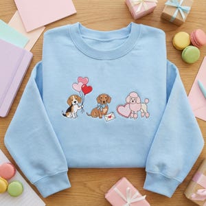 Peut inclure: Sweat-shirt bleu clair avec des broderies de chiens : un beagle avec des ballons en forme de cœur, un teckel avec un nœud et un caniche avec un cœur. Un cadeau idéal pour les amoureux des chiens.