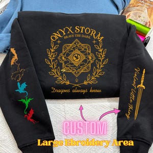 May include: Black sweatshirt with gold embroidery featuring a design with the text "ONYX STORM BRAVE THE DARK" and "Dragons always know". The sweatshirt has a large embroidery area on the front and smaller embroidery areas on the sleeves.
