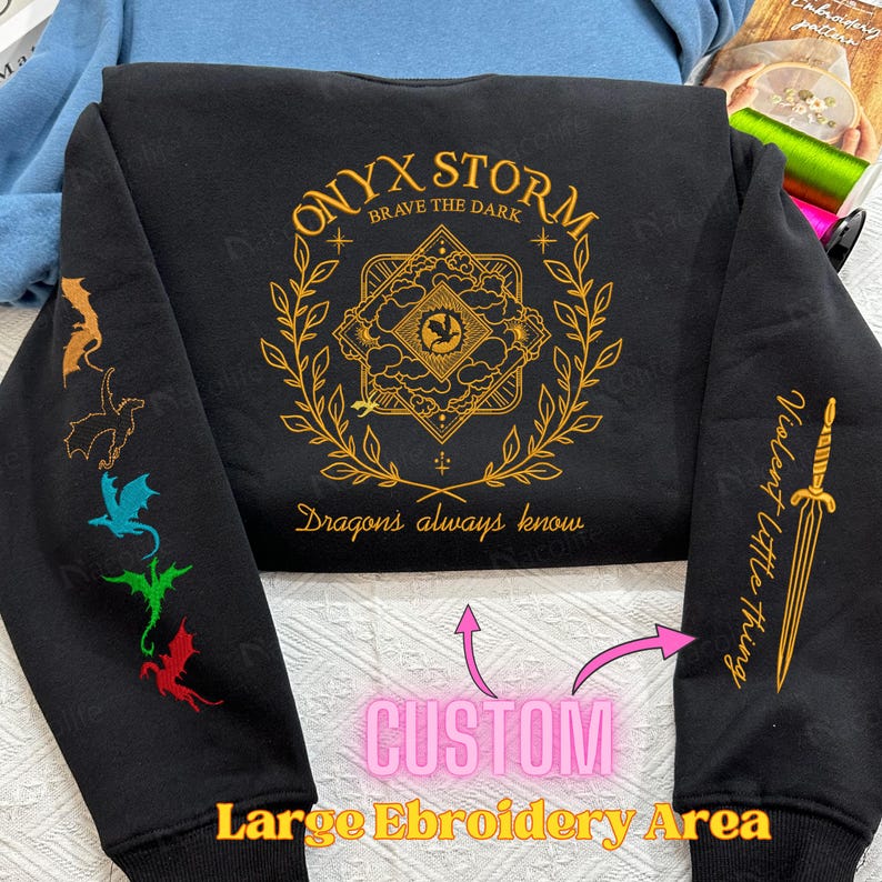 May include: Black sweatshirt with gold embroidery featuring a design with the text "ONYX STORM BRAVE THE DARK" and "Dragons always know". The sweatshirt has a large embroidery area on the front and smaller embroidery areas on the sleeves.