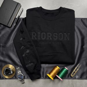 May include: A black crewneck sweatshirt featuring the embroidered text RIORSON SHADOW DADDY on the chest. The left sleeve displays a vertical sequence of five embroidered dragon silhouettes. The embroidery is in a tonal black thread.