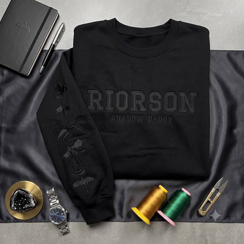 Puede incluir: Una sudadera negra de cuello redondo con el texto bordado RIORSON SHADOW DADDY en el pecho. La manga izquierda muestra una secuencia vertical de cinco siluetas de drag&oacute;n bordadas. El bordado est&aacute; realizado con hilo negro tonal.