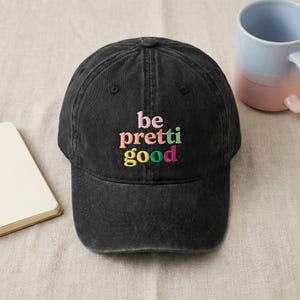 May include: A black baseball cap with a washed-out finish featuring the embroidered text "be pretti good" in multi-coloured letters. The word "be" is pink, "pretti" is in orange and green, and "good" is in yellow, green, and pink.
