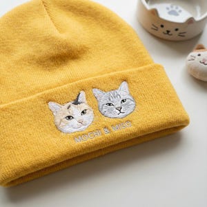 May include: Mustard yellow beanie featuring embroidered cat faces and the text "MOCHI & MISO". The beanie has a folded cuff and is made of a soft, knitted material. A cat-themed ceramic bowl and a small cat plush are in the background.