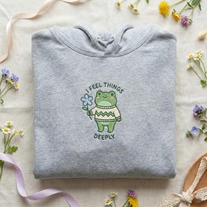 May include: A heather gray hoodie with a hood and a drawstring. The front features an embroidered frog holding a blue flower, wearing a sweater, and the text "I FEEL THINGS DEEPLY". The hoodie is surrounded by flowers and ribbons.