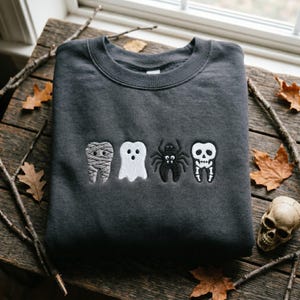 May include: A charcoal grey crewneck sweatshirt featuring four embroidered icons in a horizontal row: a tooth wrapped like a mummy, a white ghost, a black spider, and a tooth designed to look like a skull with white bone details.