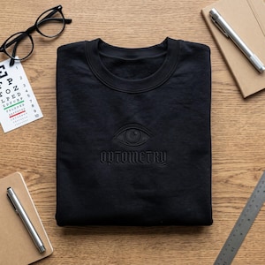 May include: A black crewneck sweatshirt featuring an embroidered design on the chest. The design consists of a detailed eye graphic positioned above the word OPTOMETRY in a stylised font. The embroidery is tonal, matching the black fabric.