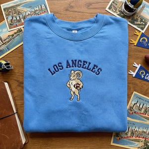May include: Light blue crewneck sweatshirt with embroidered "LOS ANGELES" text and a ram mascot holding a football. The sweatshirt is a solid light blue color.