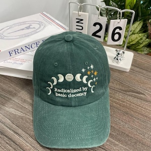 May include: Green baseball cap featuring embroidered moon phases and stars, with the text "Radicalized by basic decency" below. The cap has a curved peak and a button on top.