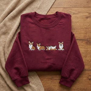 May include: A maroon sweatshirt featuring an embroidered design of four corgi dogs in various poses. The corgis are depicted in a cartoon style with brown and white markings. The sweatshirt is folded on a wooden surface.