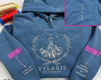 May include: A blue hooded sweatshirt with a white embroidered design of a mountain range, stars, and a sun. The text "VELARIS CITY OF STARLIGHT" is embroidered below the design. The sleeves have embroidered text that reads "to the stars who listen" and "and the dreams that are answered".