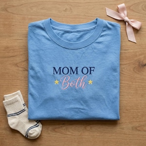 May include: A light blue t-shirt with the embroidered text "MOM OF Both" in navy and pink, with yellow stars. A pair of white socks with blue stripes and a pink bow are also present.