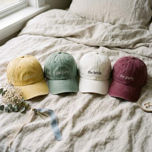 May include: Four baseball caps in yellow, green, white, and burgundy. Each cap has the text "the bride" or "the party" embroidered on the front. The caps are arranged on a bed with neutral-colored bedding.