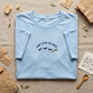 Peut inclure: Un t-shirt bleu clair à col rond avec une broderie représentant trois fourmis bleu foncé suivant une piste en pointillés vers une feuille verte et une miette brune, avec le texte CAN'T STOP THE ANTS brodé en arc au-dessus des fourmis.