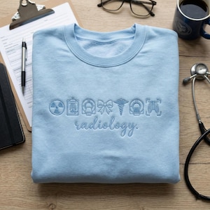 May include: A pale blue sweatshirt featuring the word "radiology" embroidered in a cursive script. Above the word are medical symbols. The sweatshirt is on a wooden surface with a stethoscope, a mug, and a notepad.