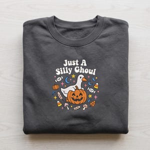Peut inclure: Un sweat-shirt gris foncé à col rond avec une impression graphique portant le texte Just A Silly Ghoul. Le motif représente une oie blanche assise sur une citrouille d'Halloween orange, entourée de petits dessins de bonbons, d'étoiles et de feuilles.
