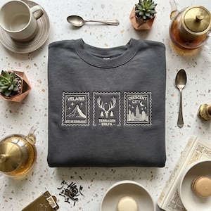 Peut inclure: Sweat-shirt gris foncé avec des motifs de timbres brodés. Les timbres représentent une montagne, un cerf et un paysage urbain, avec les textes "VELARIS CITY OF STARLIGHT", "TERRASEN - ERILFA" et "CRESCENT CITY".