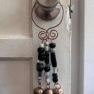 May include: A decorative hanging ornament featuring a copper wire loop with two spiral accents. Three strands of beads in black, white, and clear glass hang from the wire, each ending in a rustic, textured copper-toned metal bell at the bottom.