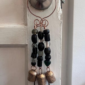 May include: A decorative hanging ornament featuring three strands of beads suspended from a copper wire ring. The beads are a mix of black, clear, and dark green shapes. At the bottom of each strand hangs a rustic, weathered brass-toned metal bell.