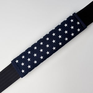 May include: A navy blue seatbelt cover with white stars. The cover is made of fabric and is designed to protect the seatbelt from wear and tear.