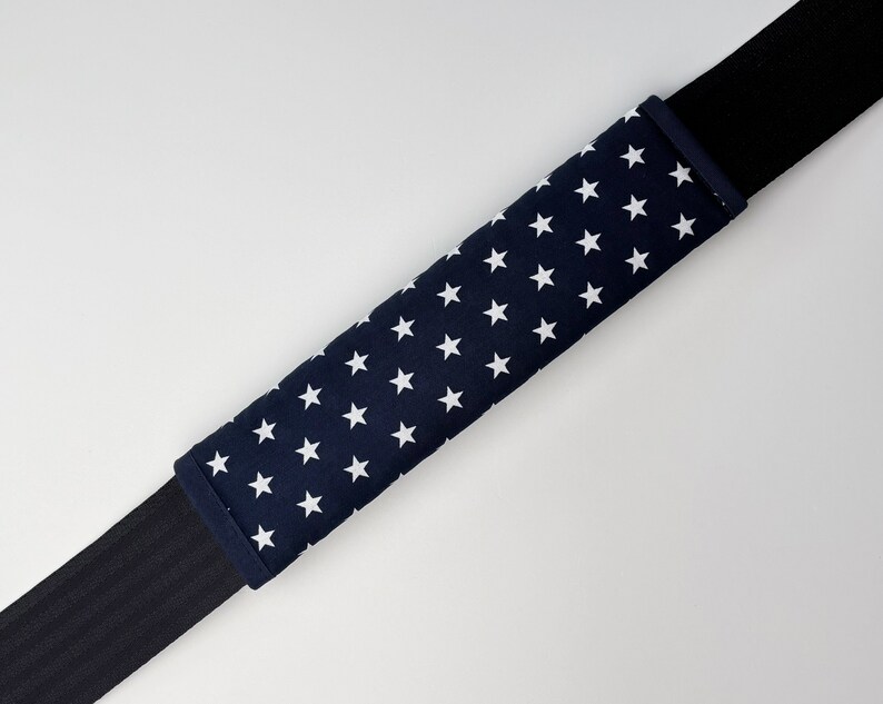 May include: A navy blue seatbelt cover with white stars. The cover is made of fabric and is designed to protect the seatbelt from wear and tear.