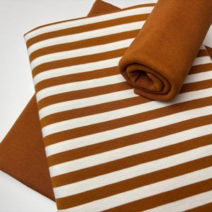 May include: A collection of fabric pieces in shades of brown and white. One piece features wide horizontal stripes, while another is a solid color. A rolled-up piece of fabric in a solid brown color is also present.