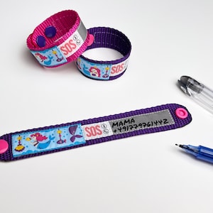 Puede incluir: Dos pulseras ajustables moradas con dise&ntilde;os de sirenas y tiras reflectantes. Una pulsera tiene una etiqueta blanca con el texto "SOS" y "MAMA +191777761472".
