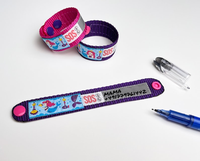Puede incluir: Dos pulseras ajustables moradas con dise&ntilde;os de sirenas y tiras reflectantes. Una pulsera tiene una etiqueta blanca con el texto "SOS" y "MAMA +191777761472".