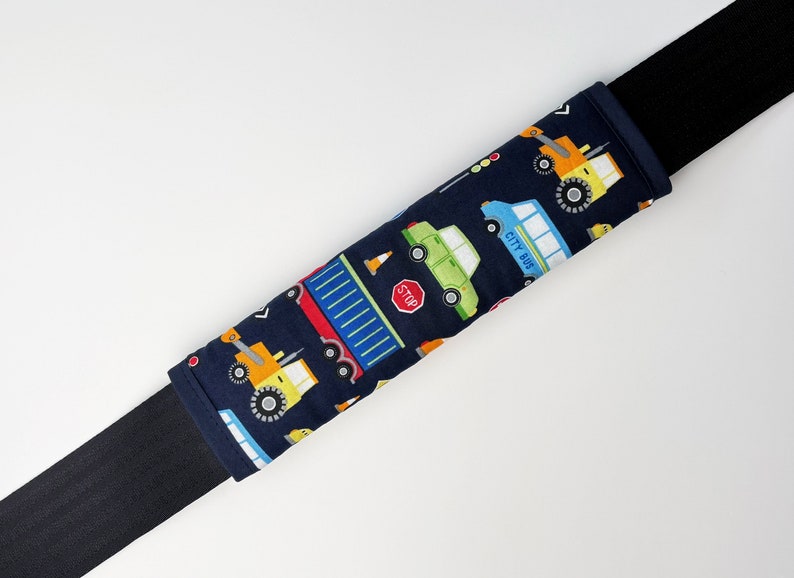May include: A blue and black car seat belt cover with a pattern of various vehicles, including a city bus, a truck, and a construction vehicle. The pattern is on a dark blue background.