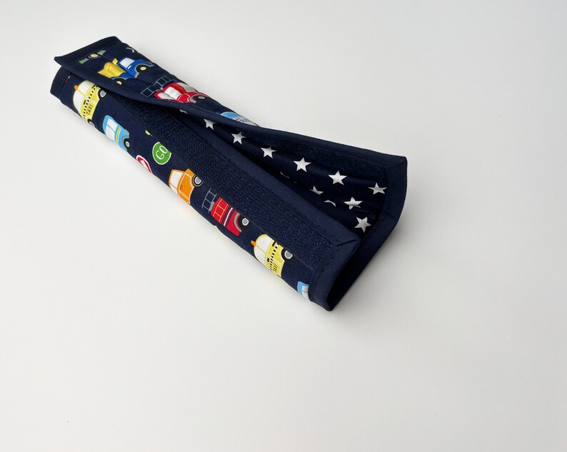 May include: A blue and white fabric car seat belt cover with a pattern of colorful cars and white stars.