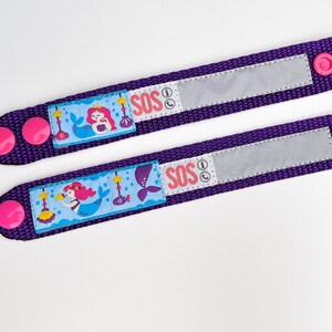 Puede incluir: Dos pulseras ajustables de color morado con botones rosas. Cada pulsera presenta un dise&ntilde;o de sirena y la palabra "SOS" con un icono de tel&eacute;fono. Las pulseras tienen un parche de velcro blanco para escribir informaci&oacute;n de contacto.