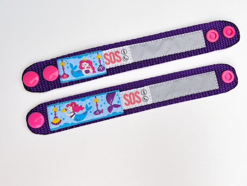 Puede incluir: Dos pulseras ajustables de color morado con botones rosas. Cada pulsera presenta un dise&ntilde;o de sirena y la palabra "SOS" con un icono de tel&eacute;fono. Las pulseras tienen un parche de velcro blanco para escribir informaci&oacute;n de contacto.