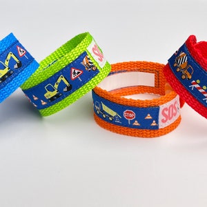Könnte beinhalten: Vier farbige Armbänder mit Baufahrzeug-Motiv und verstellbaren Verschlüssen. Die Armbänder sind blau, grün, orange und rot. Das blaue Armband hat ein Stoppschild und Baustellenkegel. Das grüne Armband hat ein Bauschild und einen gelben Bagger. Das orange Armband hat ein Stoppschild und Baustellenkegel. Das rote Armband hat einen Betonmischer und einen Kipplaster.