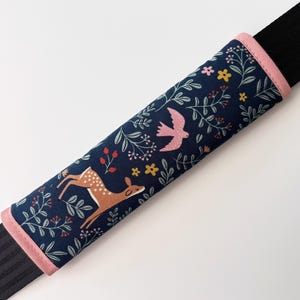 May include: A navy blue seatbelt cover with a pink trim. The fabric features a pattern of a deer, a bird, flowers, and foliage. The seatbelt cover is designed to fit over a black seatbelt.