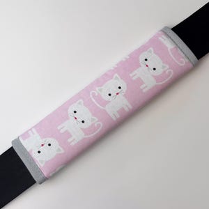 Könnte beinhalten: Ein rosa Sicherheitsgurtpolster mit einem sich wiederholenden Muster aus weißen Cartoon-Katzen. Die Katzen haben schwarze Augen und rote Nasen. Das Polster ist mit grauem Stoff eingefasst und liegt über einem schwarzen Sicherheitsgurt.