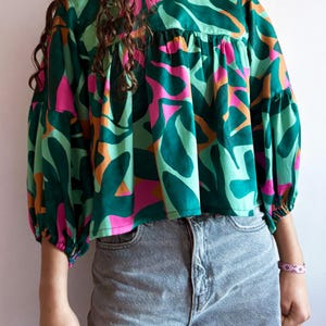 Può includere: Una blusa fantasia verde, rosa e arancione, con maniche lunghe e a sbuffo. La parte superiore ha uno scollo tondo e un dettaglio arricciato sul petto. Indossata con jeans grigi.