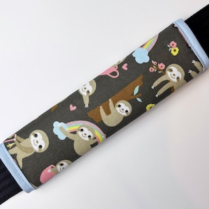 Op de afbeelding: Een blauwe en zwarte stoffen autogordelhoes met een patroon van luiaards, regenbogen en bloemen.