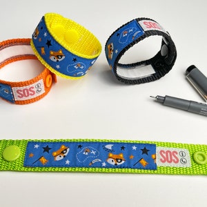 Könnte beinhalten: Mehrere verstellbare Armbänder mit einem Fuchs- und Sternmuster. Die Armbänder sind in verschiedenen Farben erhältlich, darunter Orange, Gelb und Blau. Auf einem Armband steht der Text "SOS". Ein schwarzer Stift ist ebenfalls im Bild.