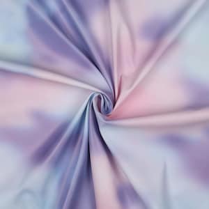 Può includere: Primo piano di un tessuto con un motivo tie-dye nei toni del viola, rosa e blu. Il tessuto è raccolto a spirale, mostrando la consistenza morbida e le transizioni di colore. L'effetto complessivo è un design onirico, simile a un acquerello.