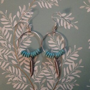 May include: A pair of silver hoop earrings with turquoise beads and a silver feather charm hanging from each hoop.