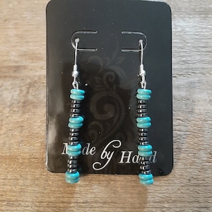 May include: A pair of dangle earrings with turquoise and black beads. The earrings are on a black card with the text "Made by Hand".