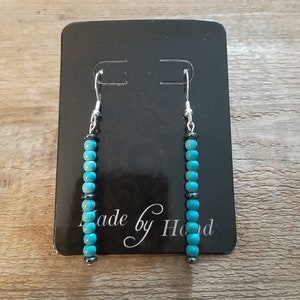 May include: A pair of turquoise and black bead earrings with silver hooks. The earrings are displayed on a black card with the text "Made by Hand".