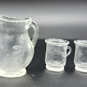May include: A set of three clear glass cups with a face design. The pitcher is larger than the two cups.