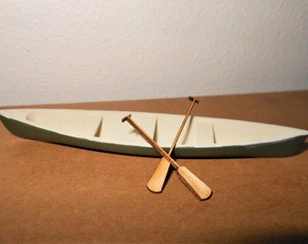May include: A miniature wooden canoe with two wooden oars. The canoe is painted green and white, and the oars are a natural wood color.