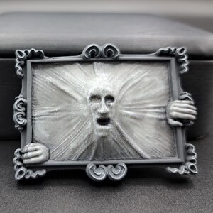 May include: A gray rectangular brooch featuring a screaming face within a decorative frame. The frame has ornate detailing and is held by two hands. The face appears to be emerging from a radiating background.