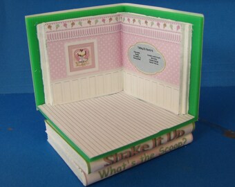 Puede incluir: Una habitación de casa de muñecas en miniatura hecha de un libro. La habitación está decorada con papel tapiz rosa y blanco, una imagen de un helado sundae y un letrero que dice "Today's Flavors". El libro se titula "Shake It Up What's the Scoop?"