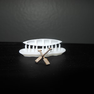 May include: A white miniature canoe with two wooden paddles. The canoe is made of plastic and has a simple design. The paddles are crossed and placed in front of the canoe.