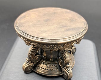Pode incluir: Um pequeno suporte de pedestal redondo na cor bronze com uma base ornamentada apresentando volutas esculpidas, motivos florais e pilares verticais. A superfície superior é plana e circular com acabamento texturizado, apoiada sobre uma base decorativa.