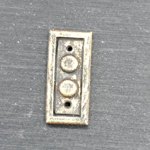 May include: A rectangular, antique-style metal plate with two circular buttons and two holes. The plate has a textured, aged appearance with a bronze-like finish. The buttons have a detailed, embossed design.