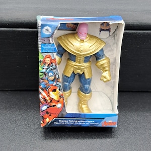 May include: A Thanos Talking Action Figure from the Avengers series. The figure is purple-skinned with a gold chest plate and blue pants. The figure is holding a purple gauntlet with gold accents. The figure is packaged in a clear plastic box with a white background and a black and red Avengers logo.