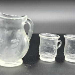 Puede incluir: Un juego de tres tazas de vidrio transparente y una jarra con un diseño de cara. La jarra tiene un asa y una base ancha. Las tazas tienen un asa y una base redonda.